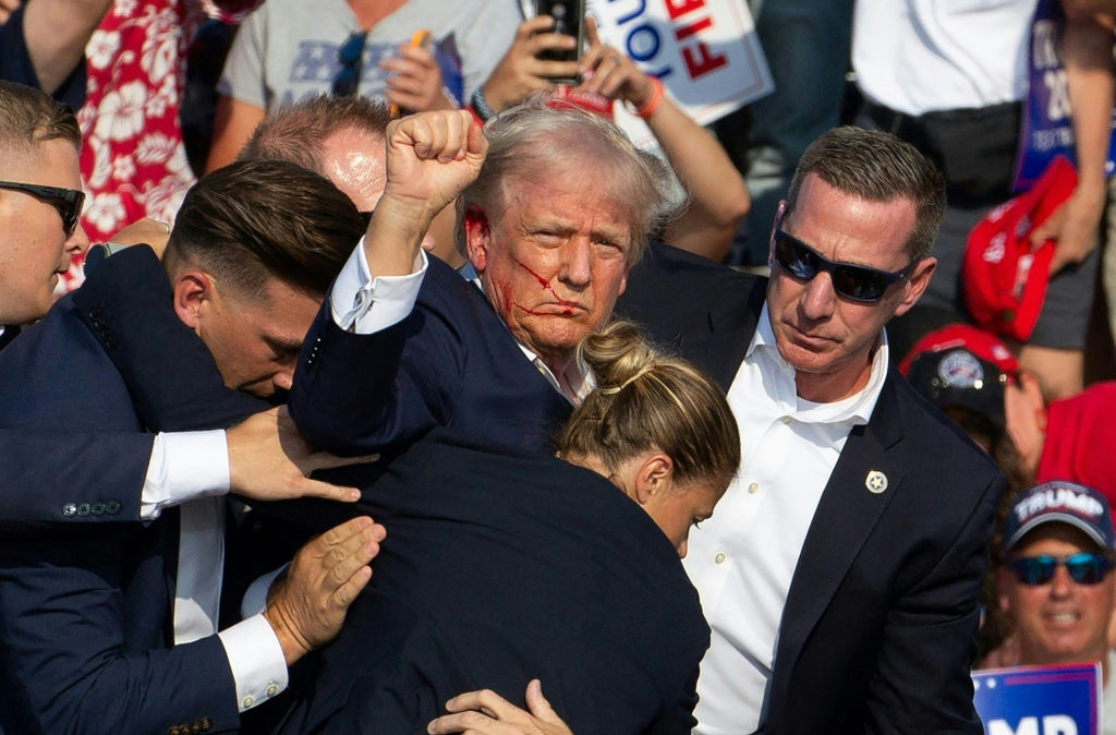 A public figure surrounded by security personnel in a crowded outdoor event, appearing serious with a clenched fist raised