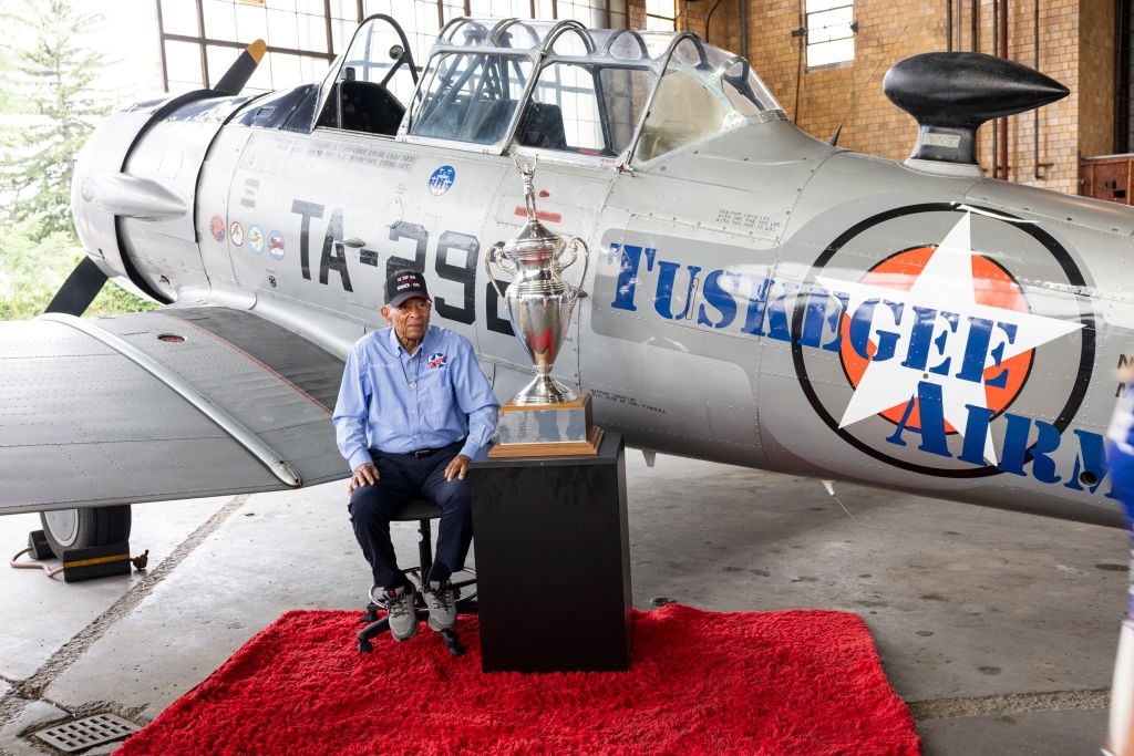 Man sitting in front of a Tuskegee Airmen plane, on a red carpet, next to a large trophy in a hangar
