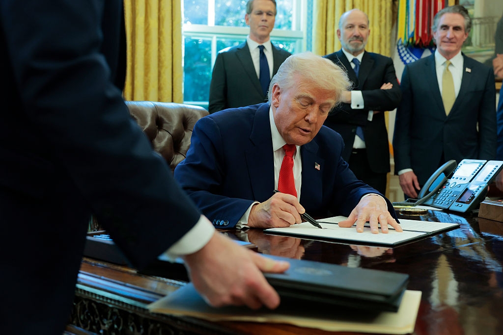 A person in a suit sits at a desk, signing a document, surrounded by others standing