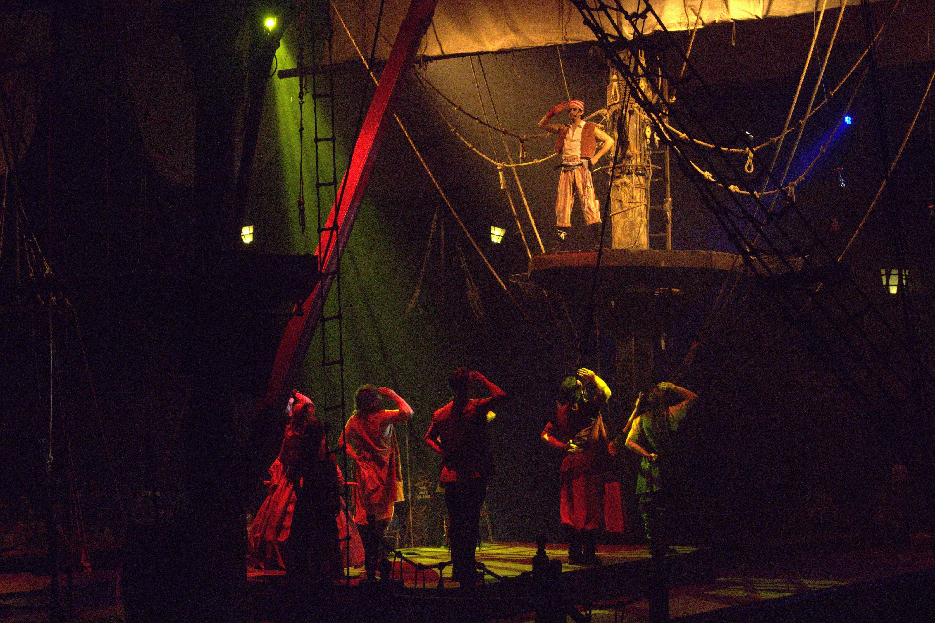 Escena teatral de piratas en un barco simulado; cinco personajes en el escenario y uno principal en posición de mando bajo foco