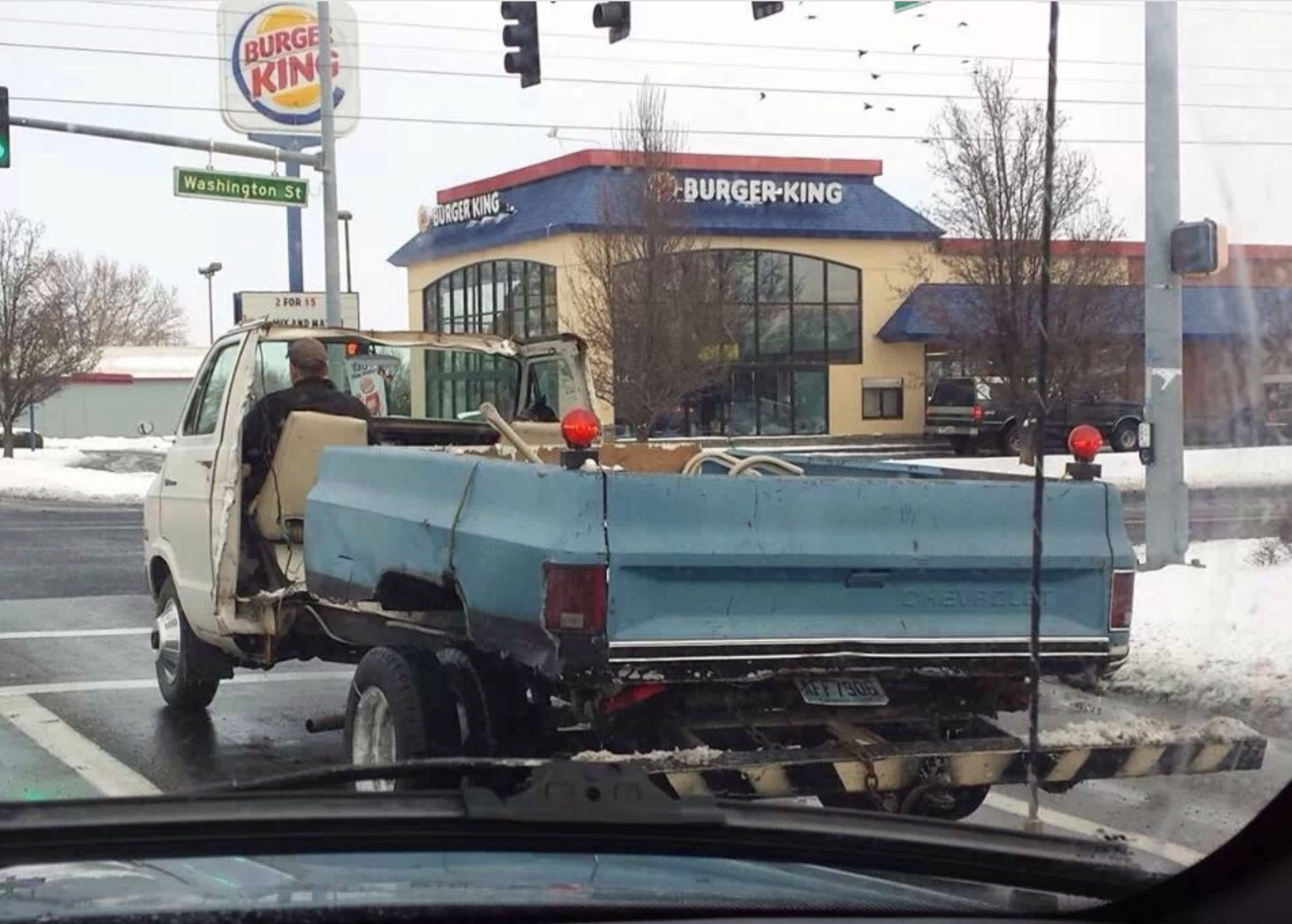 A pickup truck with a damaged, misshapen bed is stopped at an intersection near a Burger King. Snow is visible on the ground