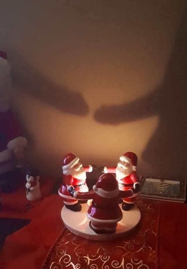 Three Santa figurines are arranged in a circle, casting an eerie shadow on the wall resembling a face. A snowman figure is nearby