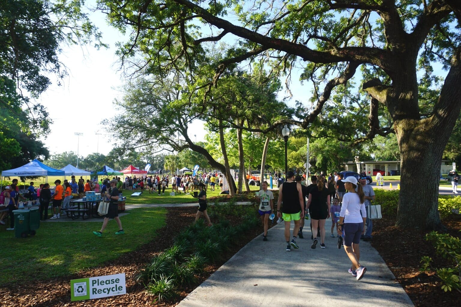 Personas caminando en un parque, rodeadas de árboles y puestos de información. Un cartel indica "Por favor, recicla". Hay un evento al aire libre