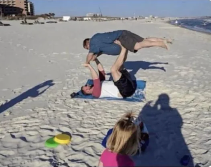 Two people perform an acroyoga pose on a sandy beach while another person takes a photo. Nearby, a child plays with beach toys