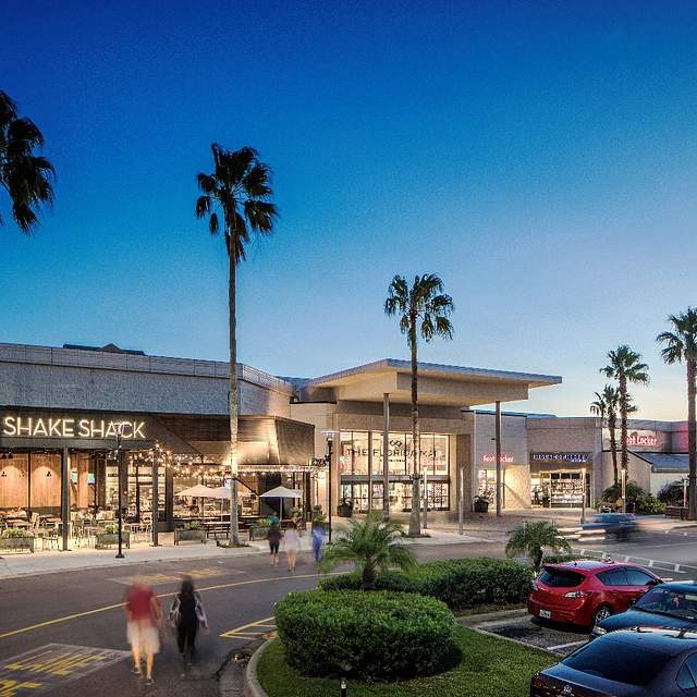 Centro comercial al aire libre con tiendas y restaurantes, rodeado de palmeras y coches estacionados. Personas caminan por el área