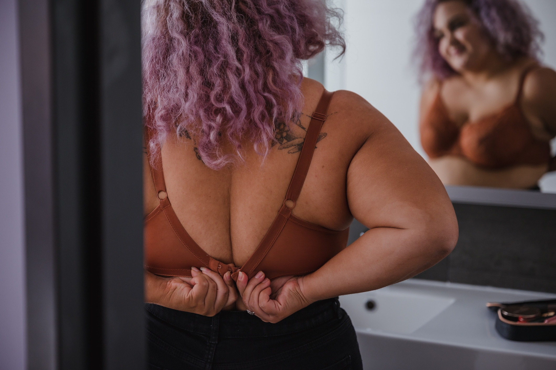 Person with curly hair shown from the back, adjusting their bra in front of a bathroom mirror, reflection partially visible