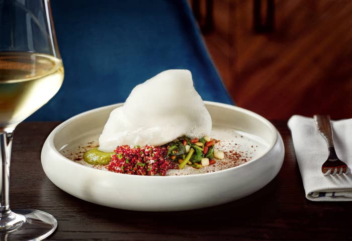 Plato gourmet con espuma blanca, quinoa roja y vegetales, junto a una copa de vino blanco sobre la mesa