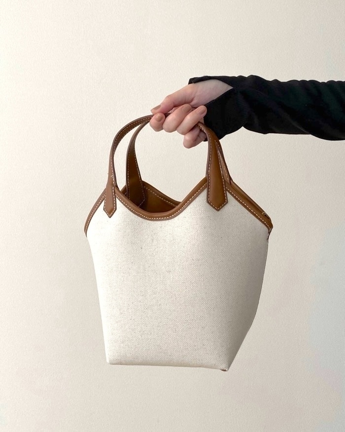 シンプルなハンドバッグを持つ黒い服の人物の手。バッグは布製で、革の取っ手が付いている。