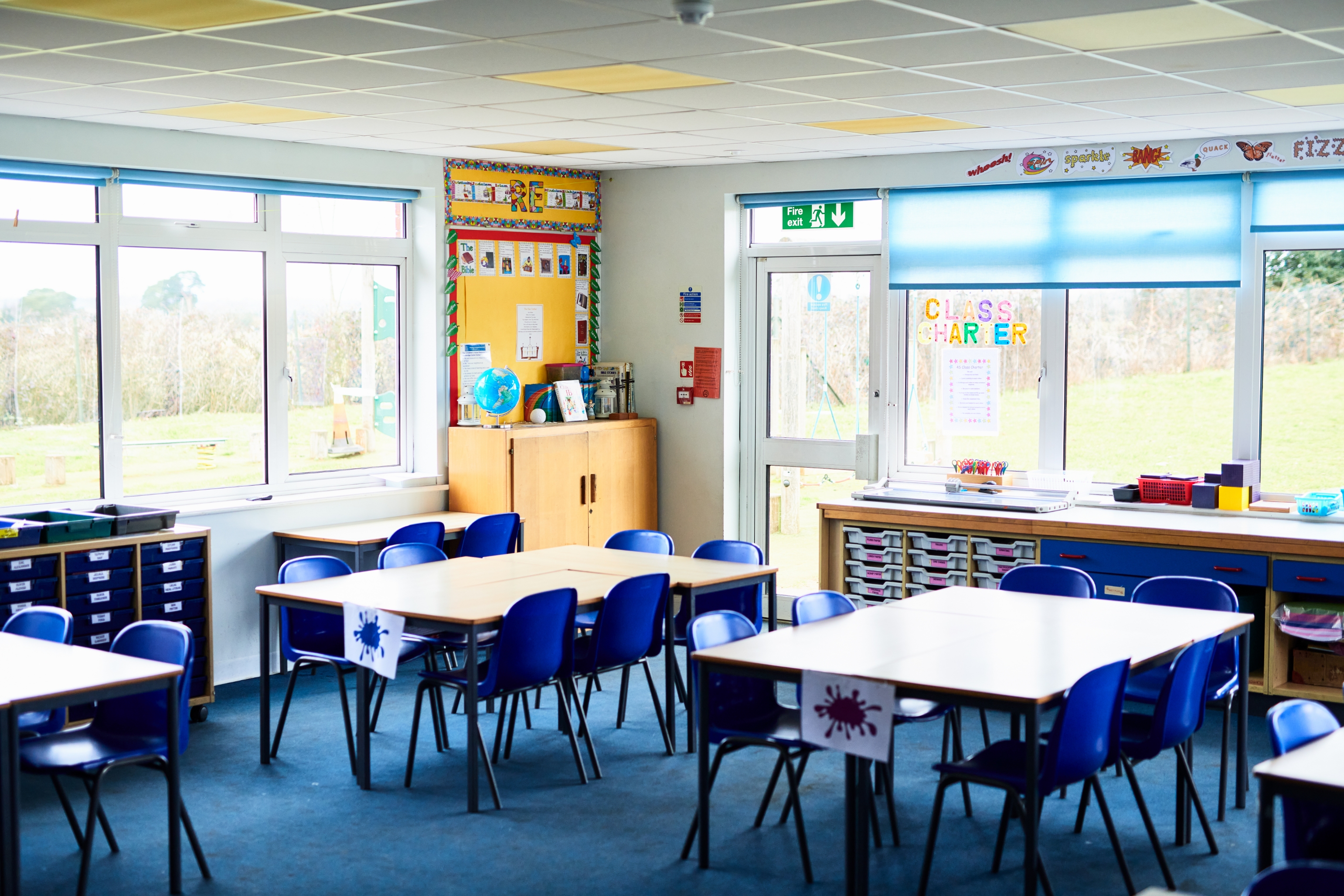 Empty classroom with blue chairs and white tables arranged in groups. Large windows overlook a grassy field. Bright, organized setting