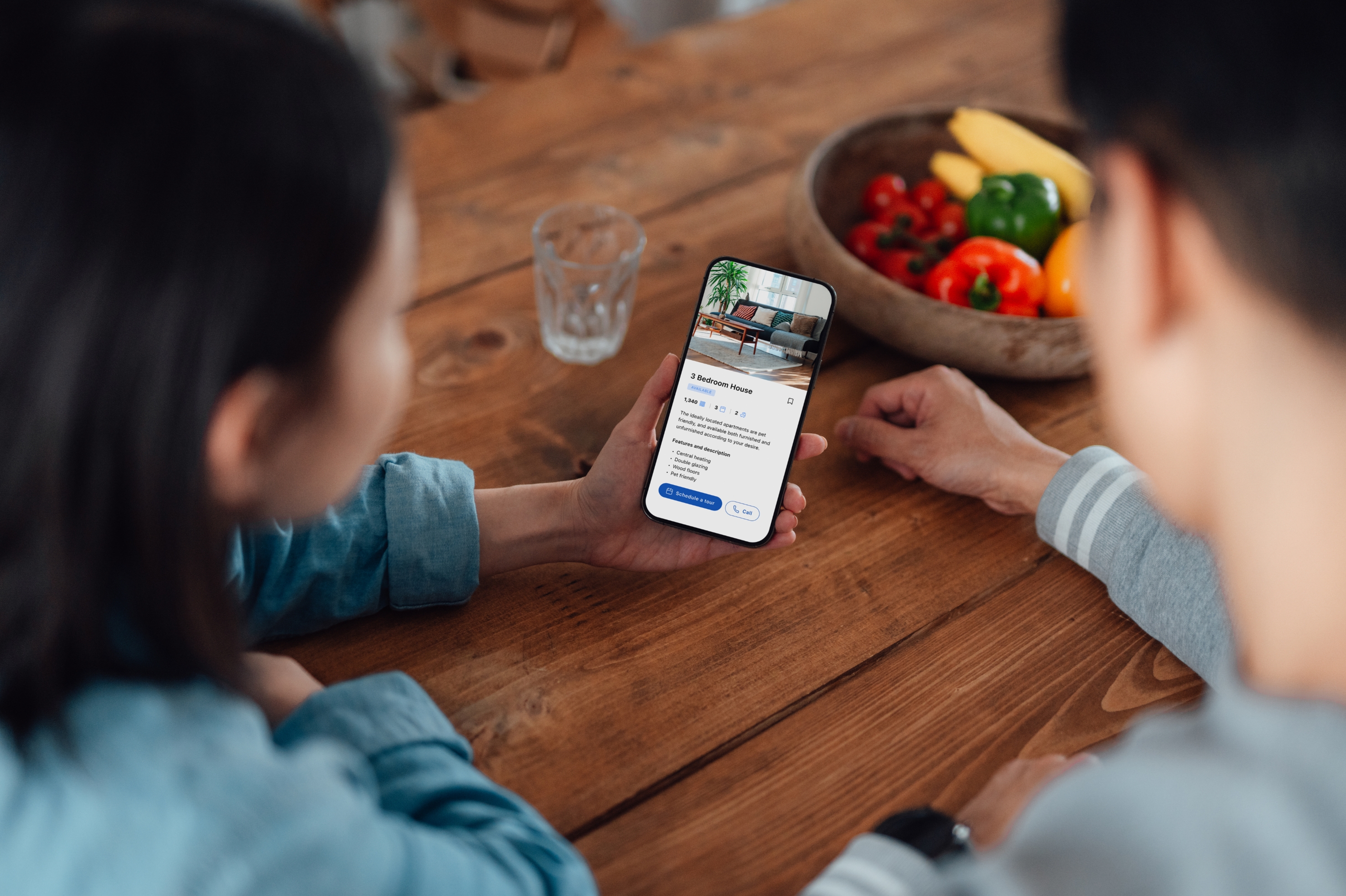 Two people sit at a wooden table looking at a smartphone screen displaying a home rental or booking app, with a fruit bowl in the background