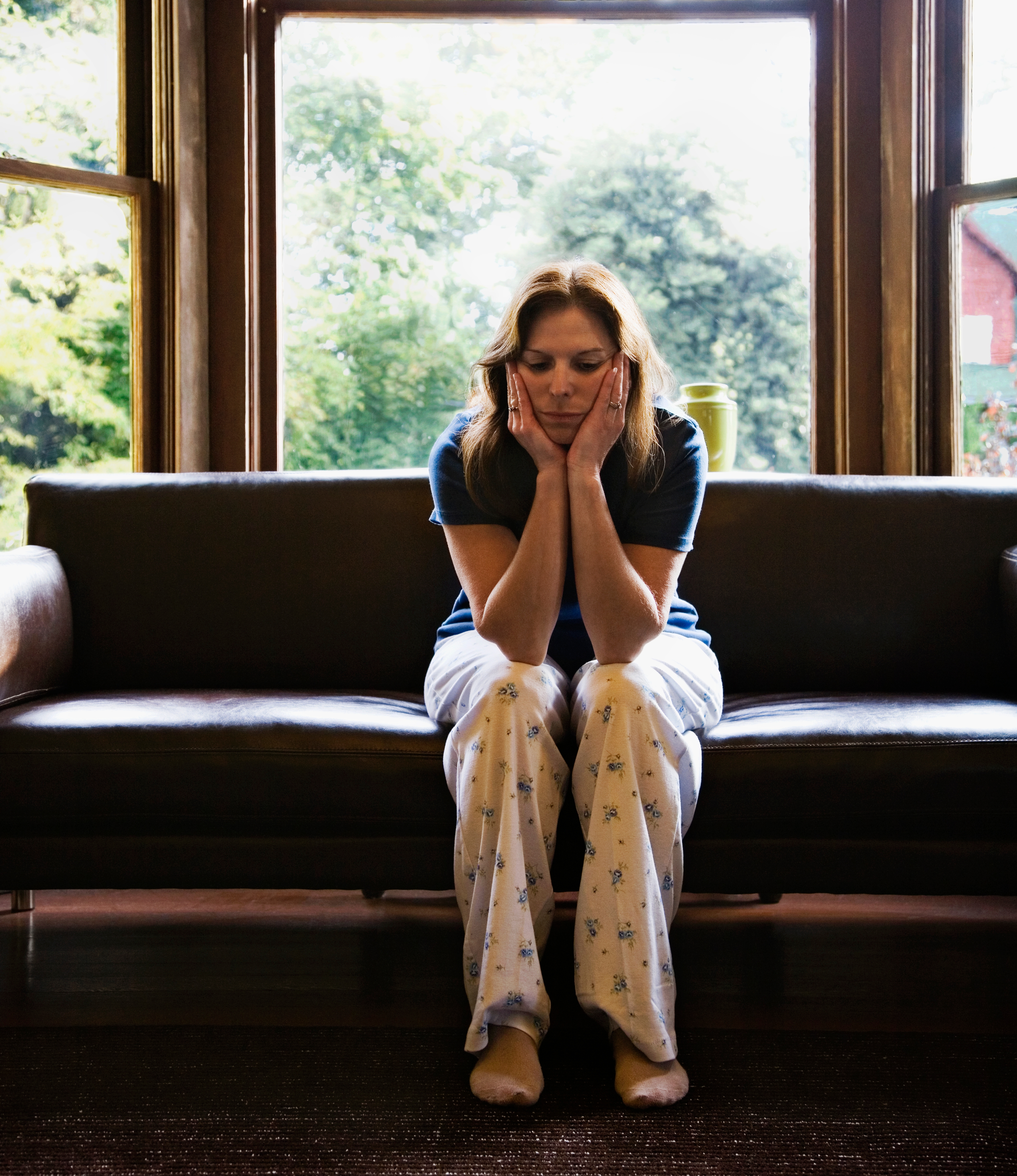 Person sitting on a couch with head in hands, wearing comfortable pajama pants, looking contemplative. Large windows reveal a leafy view outside