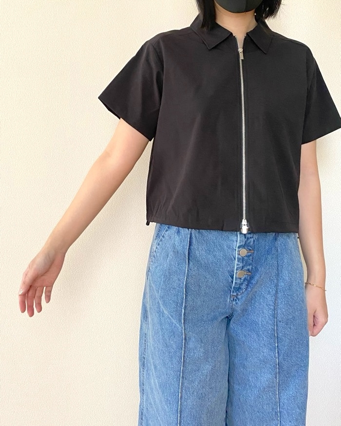 Person wearing a short-sleeved, zippered top with wide-leg jeans, standing with one arm outstretched