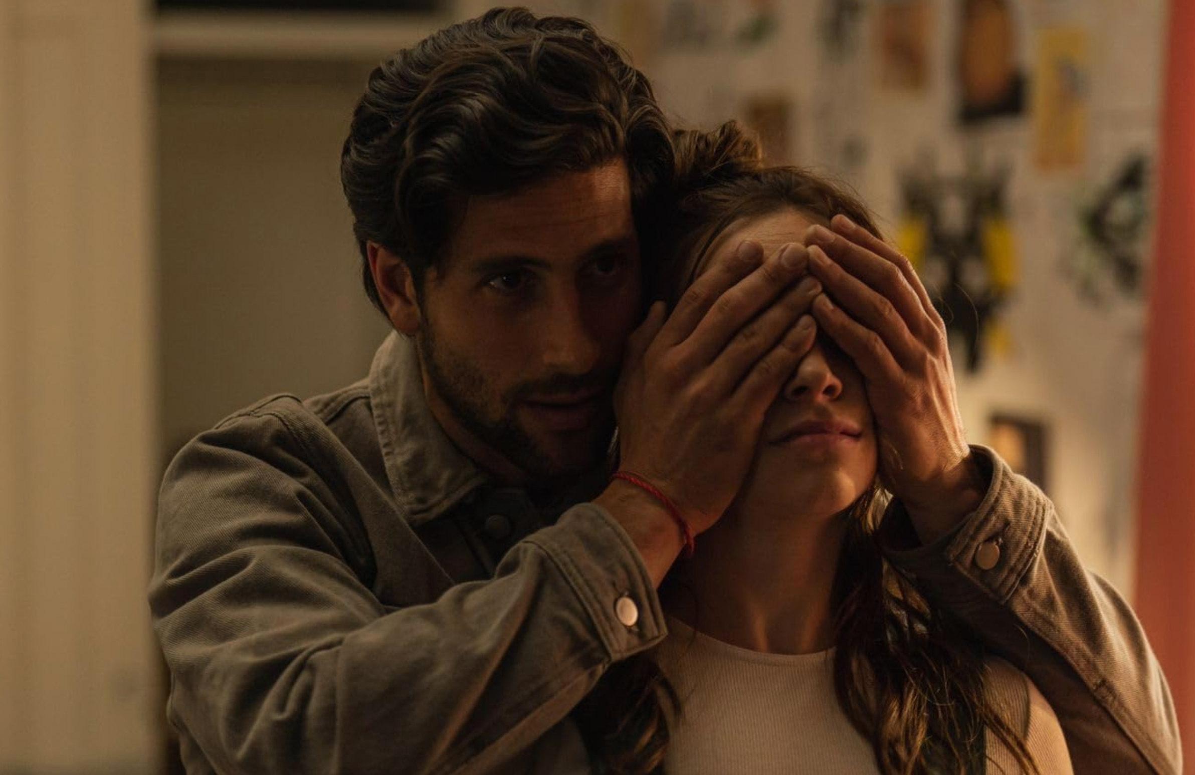 Un hombre cubre los ojos de una mujer en una escena íntima y misteriosa. Están en un ambiente interior con decoraciones de pared al fondo