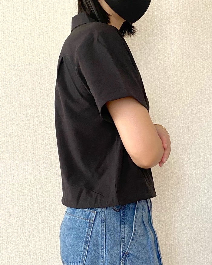 Person in casual black shirt and jeans, wearing a mask, stands with arms crossed, facing sideways