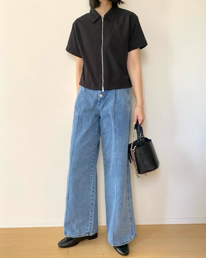 Person standing against a plain background, wearing a short-sleeve zip-up top, wide-leg jeans, and holding a textured handbag