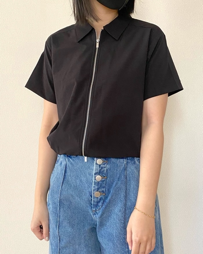 A person in a short-sleeve, zip-up black shirt and high-waisted blue jeans with large buttons, wearing a mask