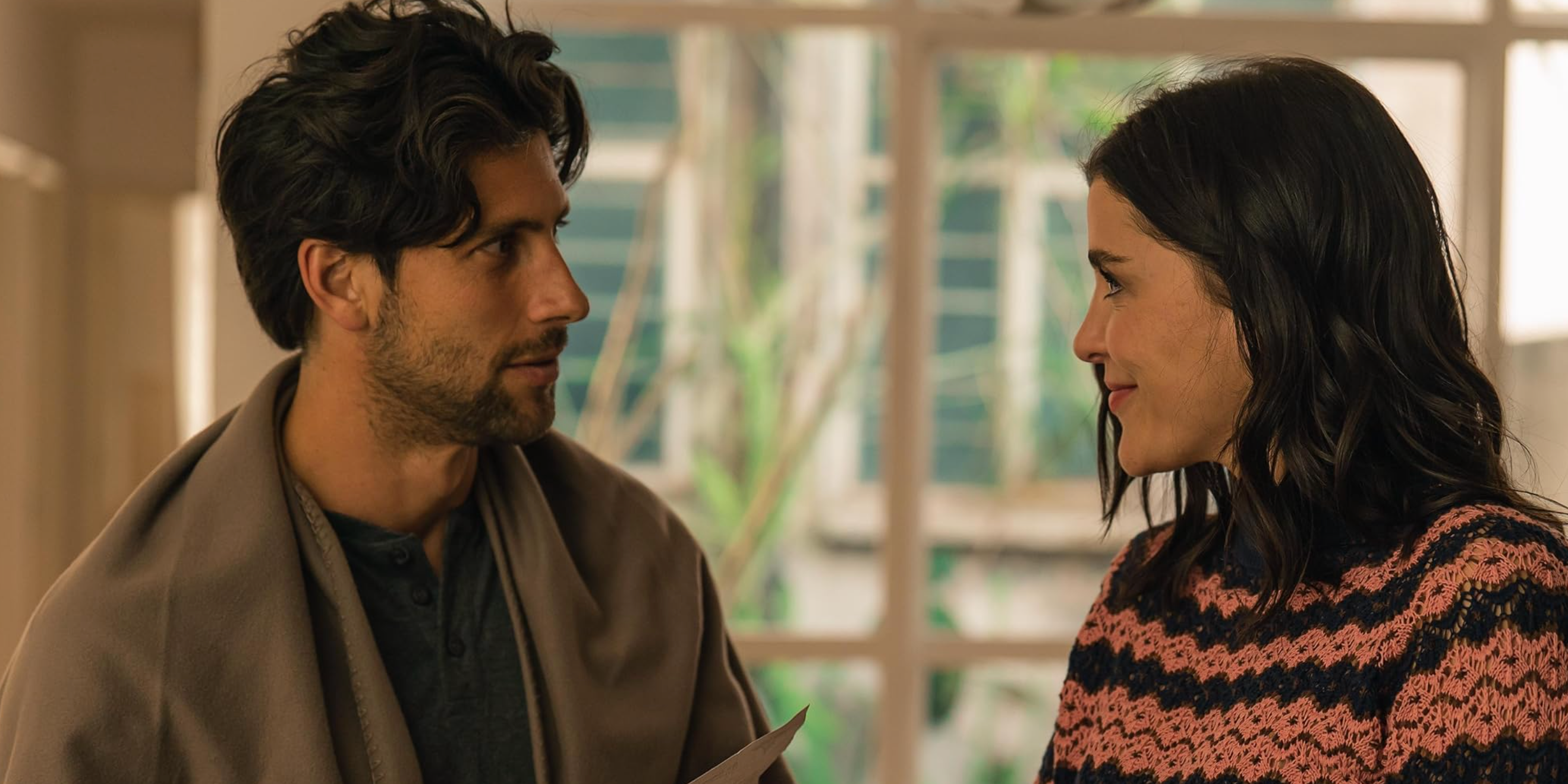 Un hombre y una mujer mirándose en un interior moderno. Él lleva una capa, y ella un suéter con patrón. Escena de película o serie