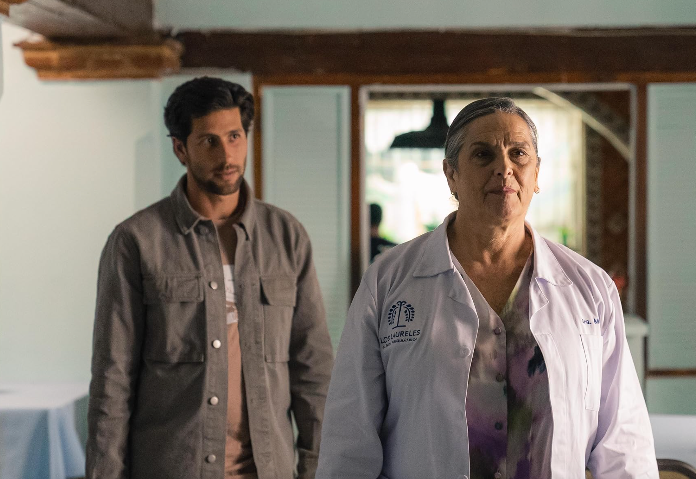 Un hombre y una mujer mayor en bata de laboratorio dentro de un edificio, escena de una serie de TV o película