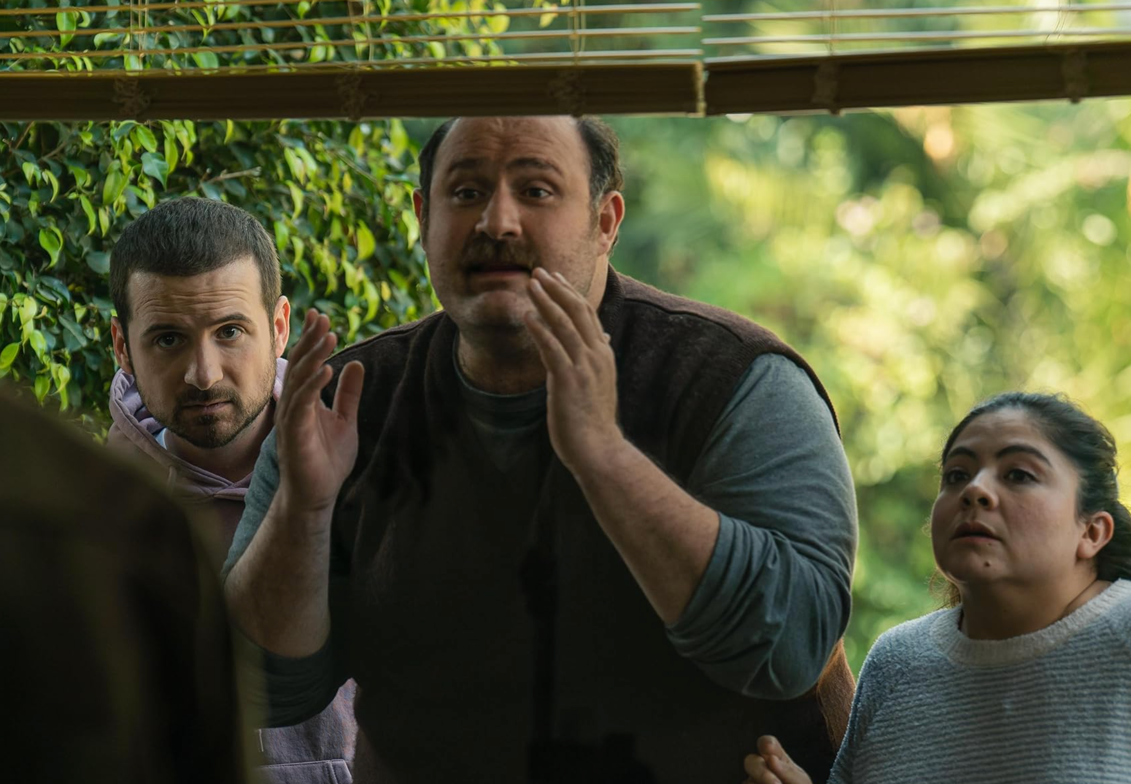 Tres personas frente a una puerta de vidrio, con expresiones preocupadas. Una parece estar hablando gestualmente. Escenario de película o serie