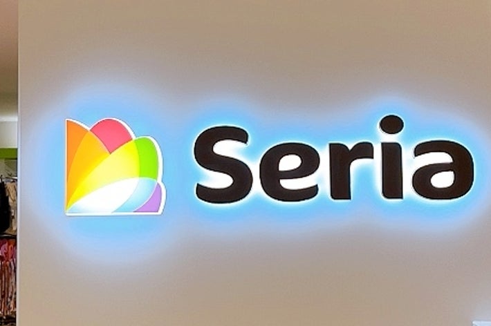 セリア店舗の内部、壁にロゴが表示されています。背景に商品棚が見えます。