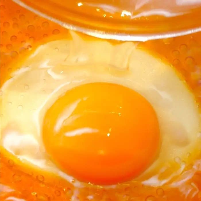 卵がフライパンで焼かれ始める様子をクローズアップで捉えた画像。卵黄は中央にあり、ジューという音が聞こえてきそうな場面。