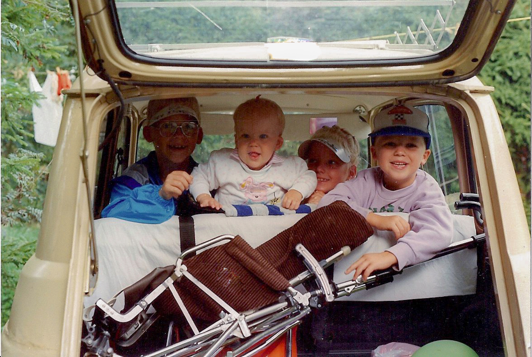 A family trip photo showing three kids and an adult sitting in an open car trunk, smiling with a stroller and balloons visible