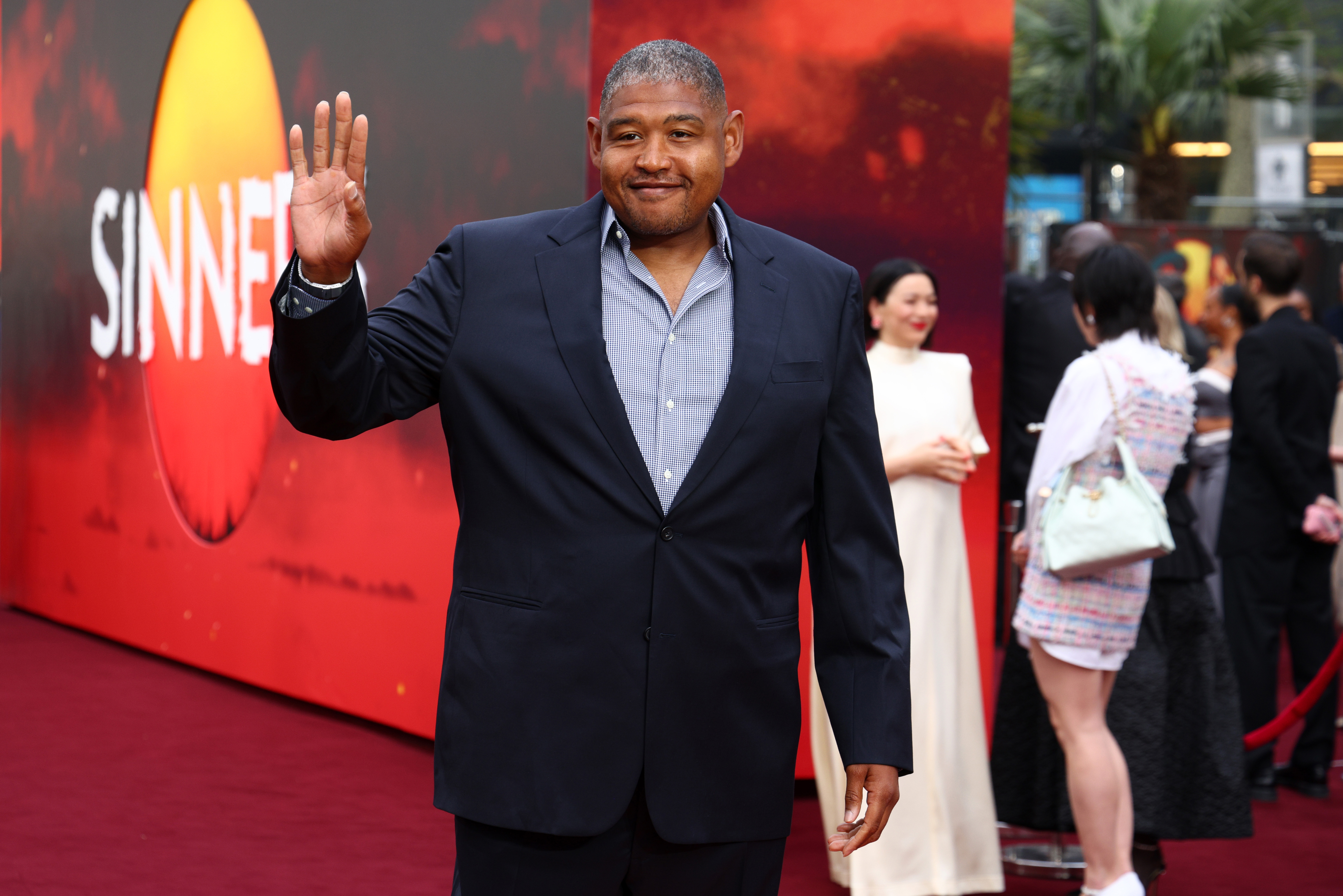 Person in a dark suit waves on a red carpet at a public event. People and a backdrop are in the background