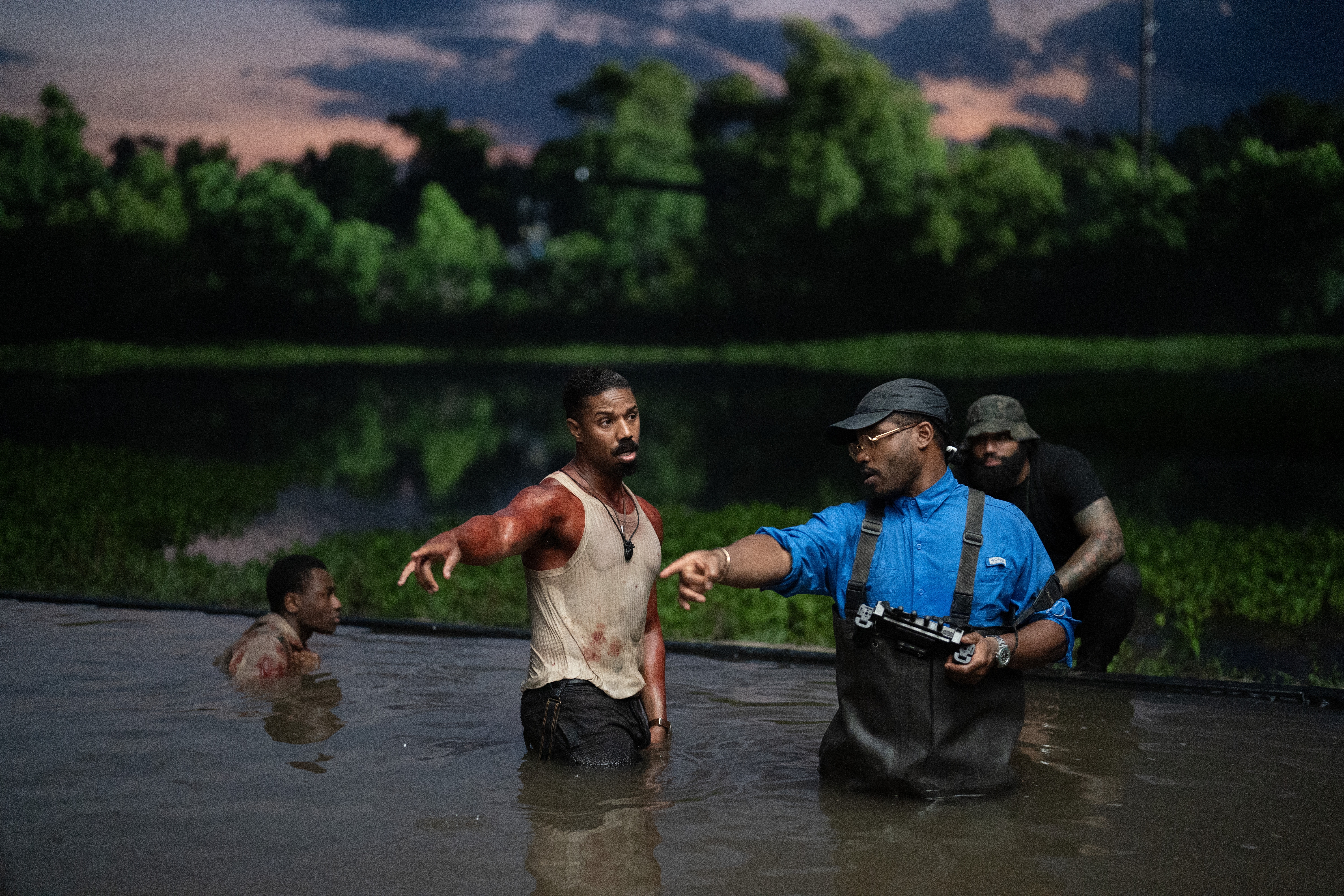 Actors in a swampy water scene, with one wearing muddy, distressed clothing, and another in waders with film equipment, talking