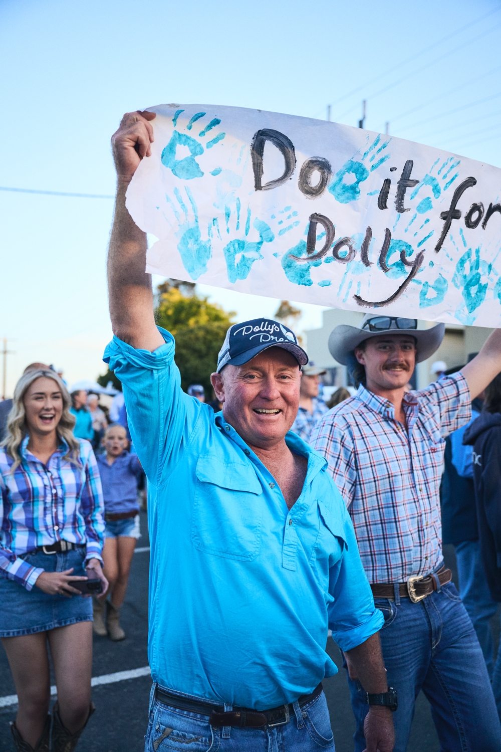 A group of people at a rally, with a man in a cap holding a &quot;Do it for Dolly&quot; sign. Others wear casual attire, smiling and supportive