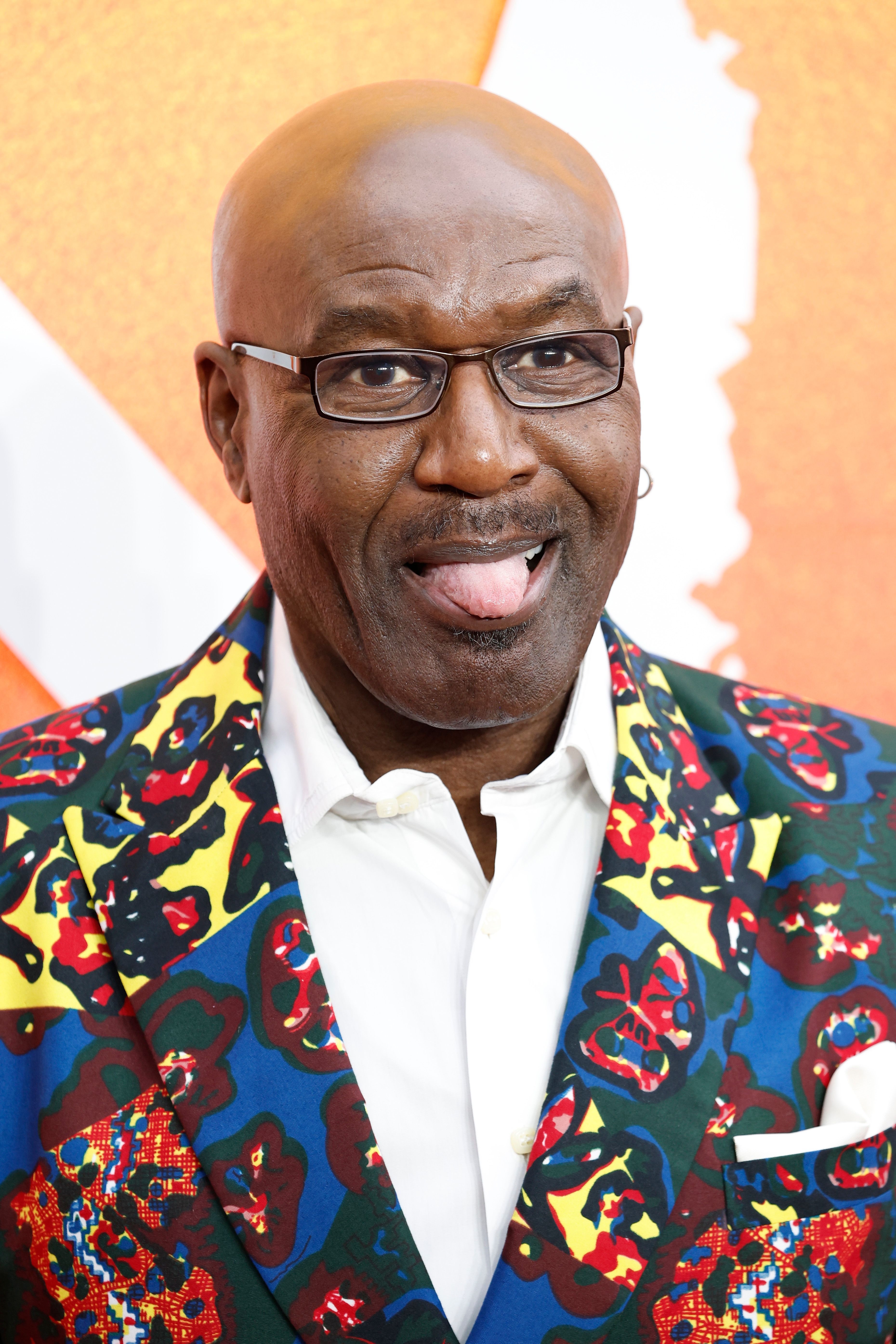Man in a patterned blazer and glasses sticks out his tongue playfully on the red carpet