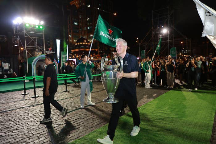 A person is walking on a green carpet holding a large trophy, surrounded by people and event lighting at night. They are wearing a polo shirt