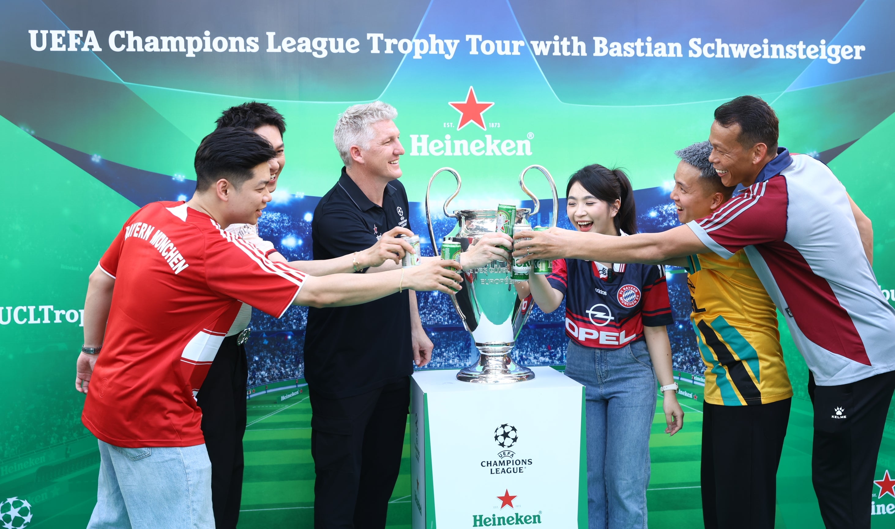 Group posing with UEFA Champions League trophy on display, featuring Bastian Schweinsteiger and fans wearing various soccer jerseys