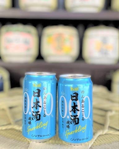 2本の青い缶に入ったスパークリングノンアルコール日本酒が正面に並んでいる。後ろに樽が複数見える。