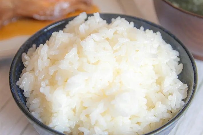 テーブルに盛られた白ご飯のアップ。背景に焼き魚とわかめの味噌汁が添えられている。