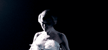 Person in a feathered costume, reminiscent of a swan or ballet attire, stands dramatically under spotlight on stage