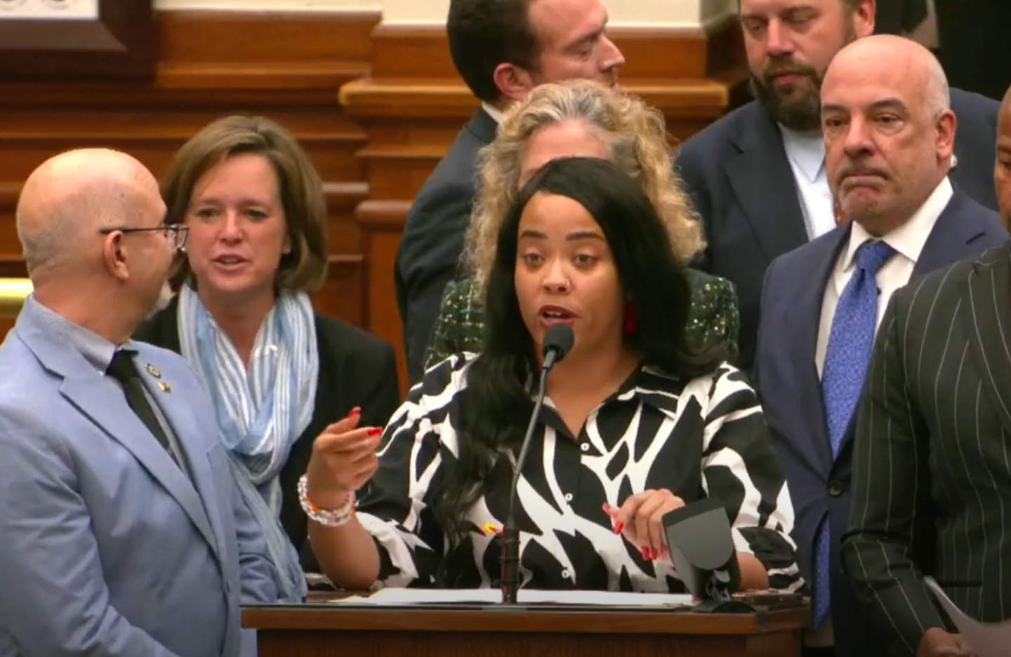 A woman speaks at a podium, surrounded by people in formal attire