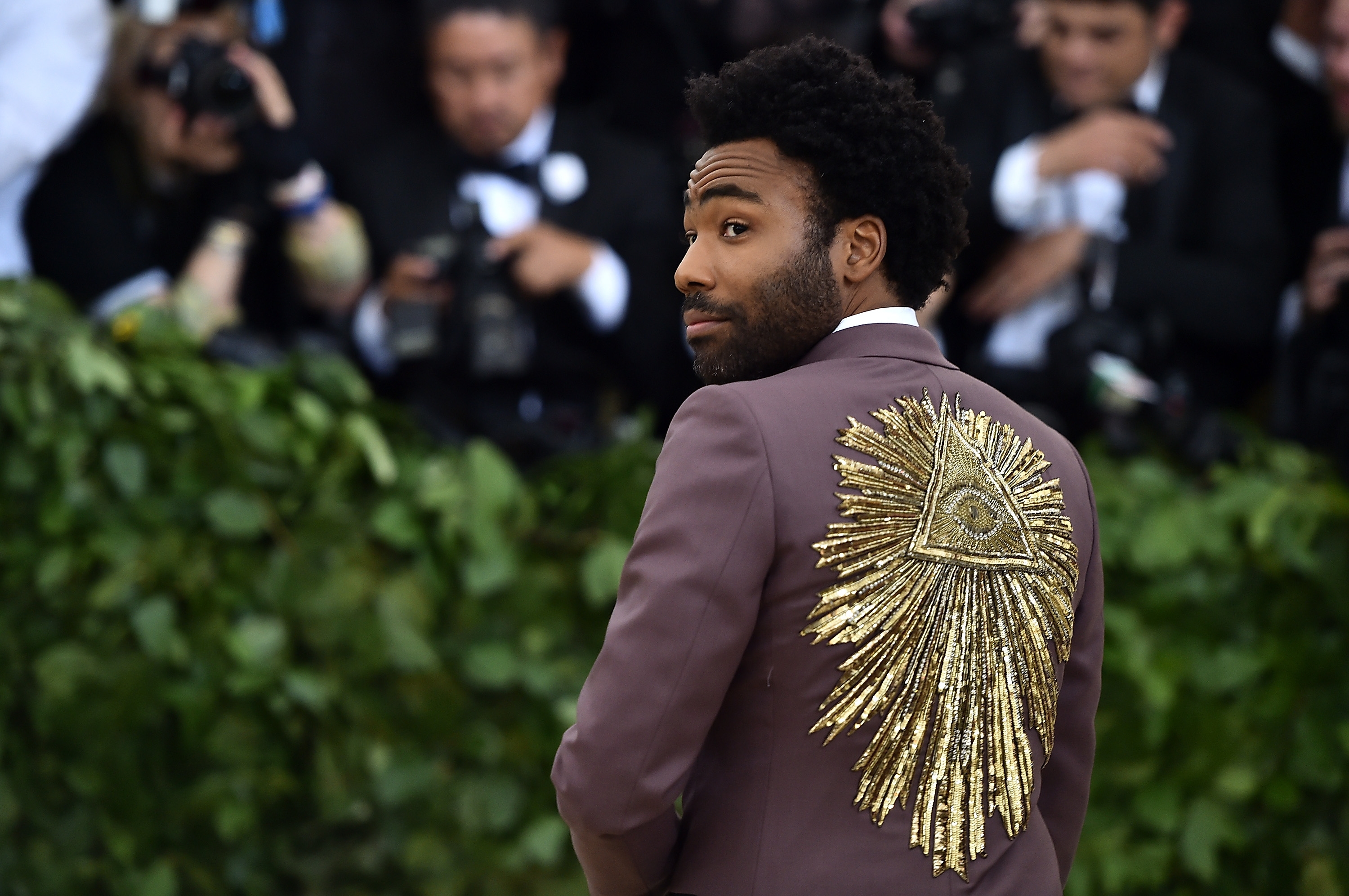 A person wearing a suit with an intricate sunburst and triangle design on the back poses for photos at a formal event