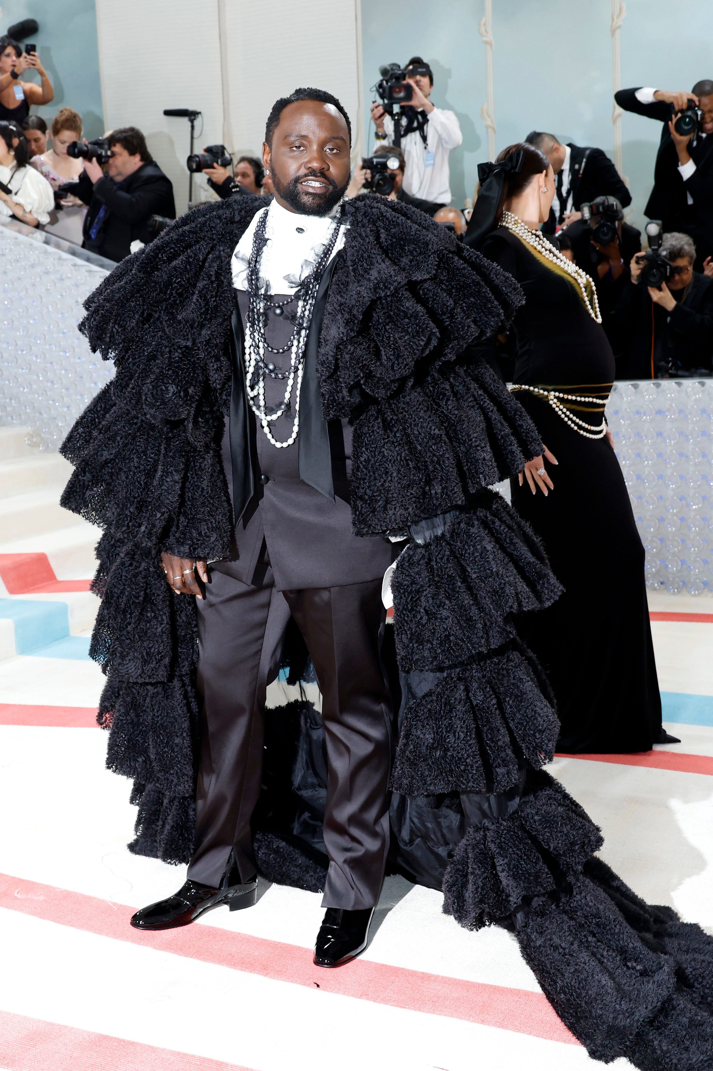 Person wearing an elaborate, ruffled black cape with layers, over a tuxedo and pearl accessories on a red carpet, surrounded by photographers