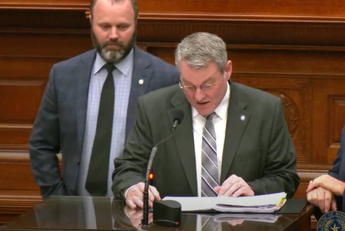 Two men stand at a podium. One reads from papers while the other observes. Formal setting with wood paneling in the background