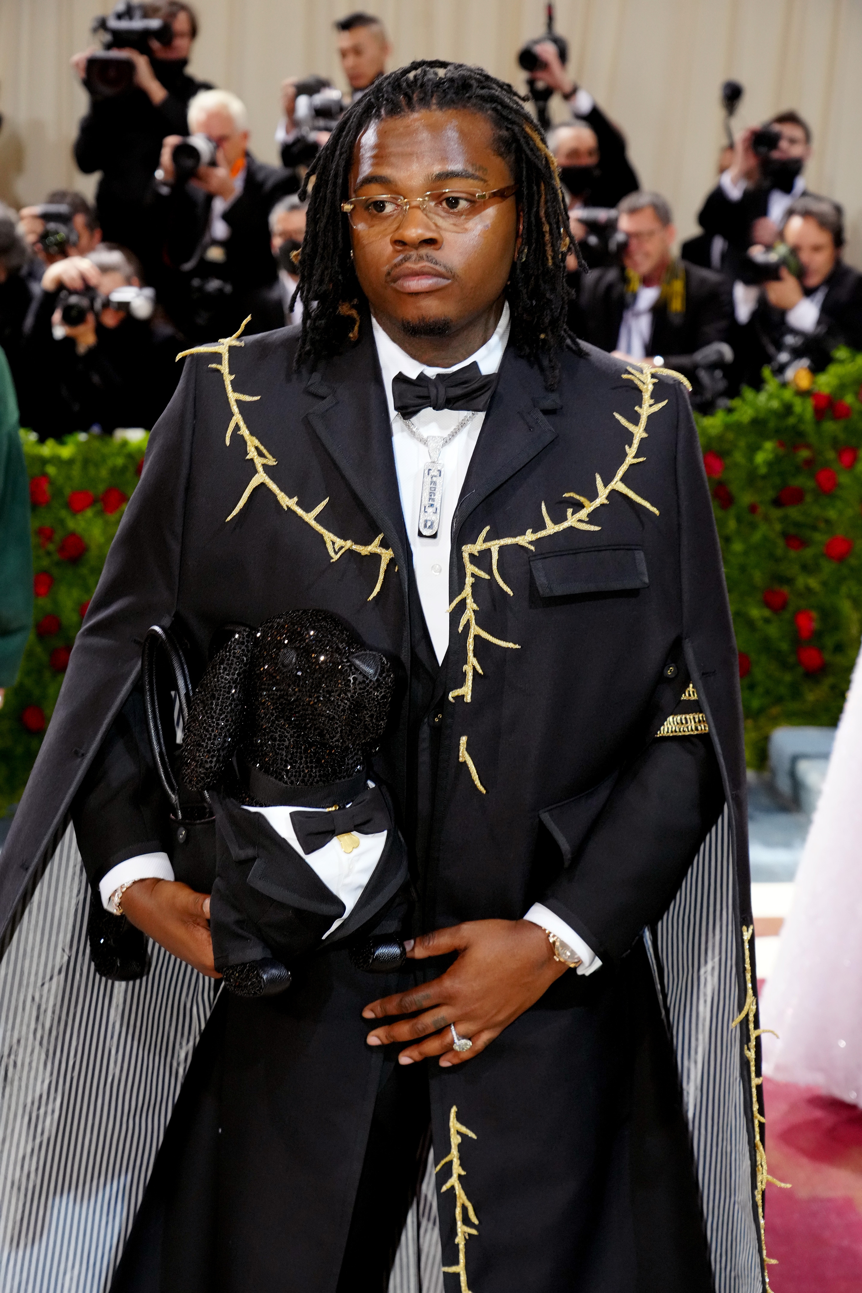 Person in a tailored suit with unique stitching details, holding a shiny accessory. Photographers are visible in the background at an event