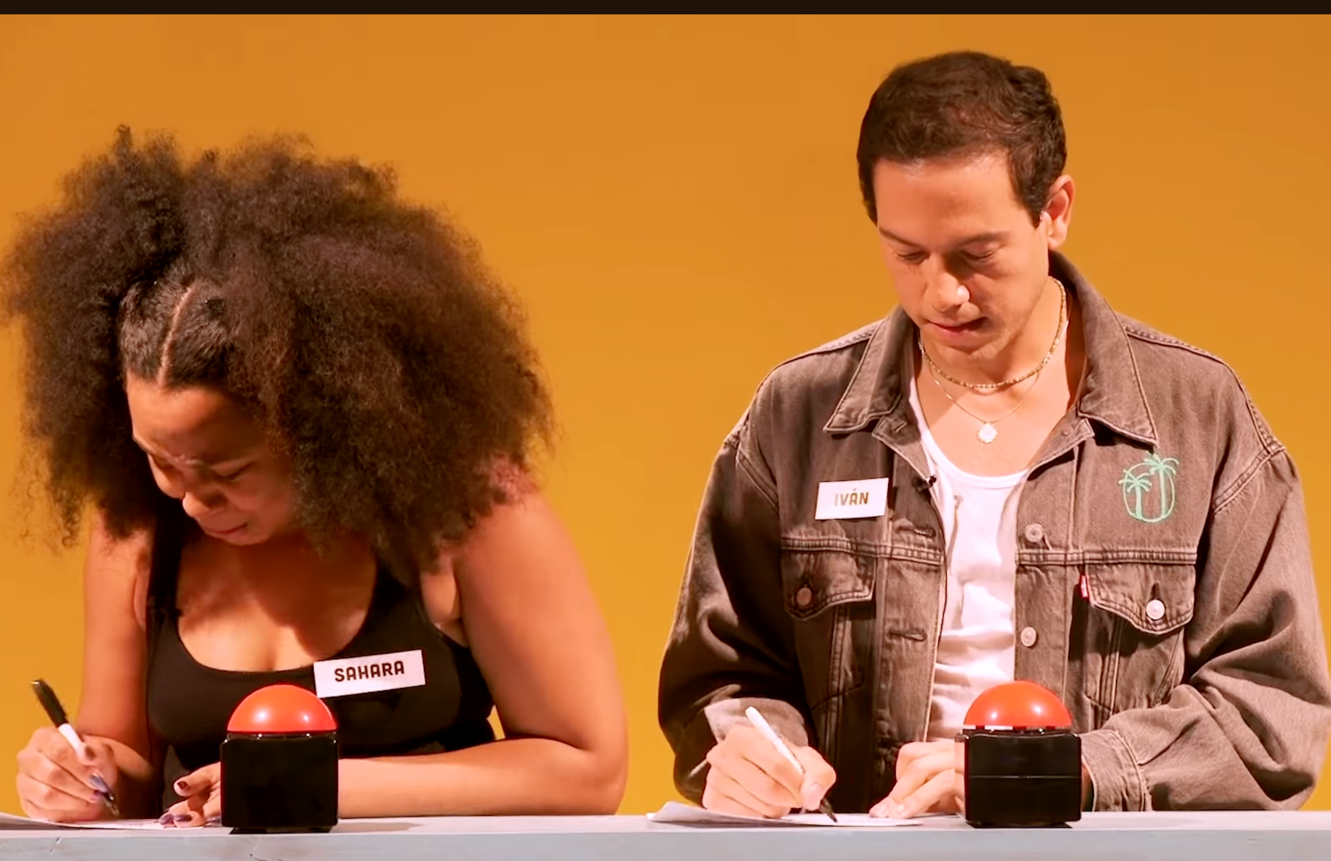 Two people sit at a table with red buzzers, concentrating on writing in what appears to be a game or quiz setting