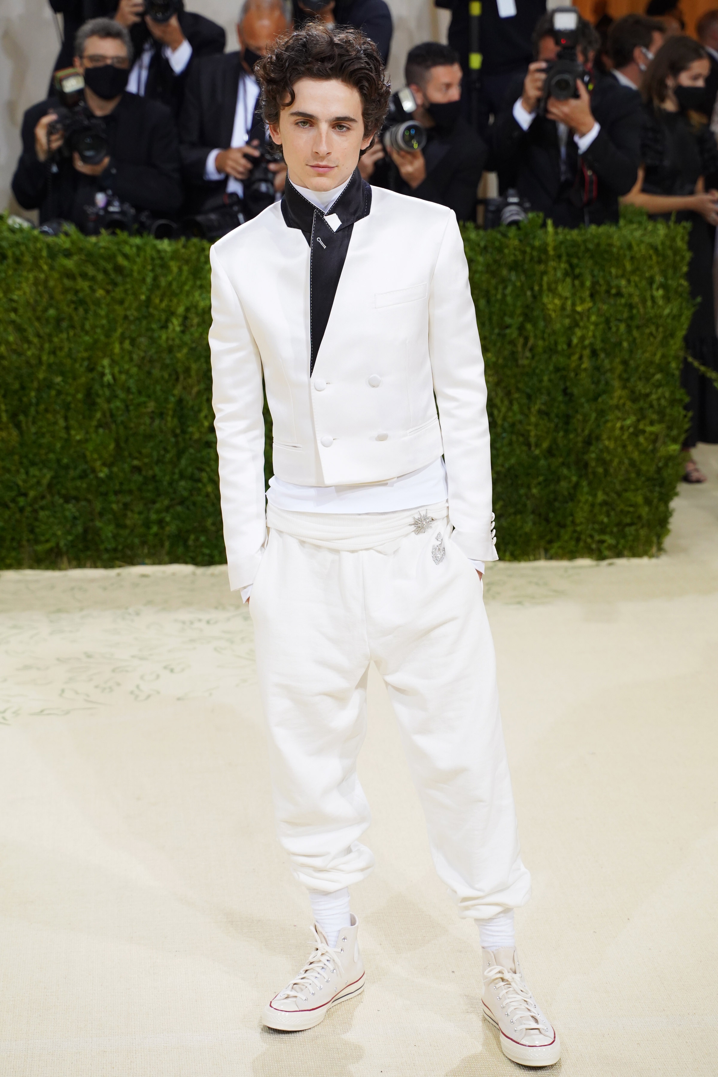 Person in a formal white suit with casual jogger pants and sneakers, standing on a red carpet. Photographers are in the background