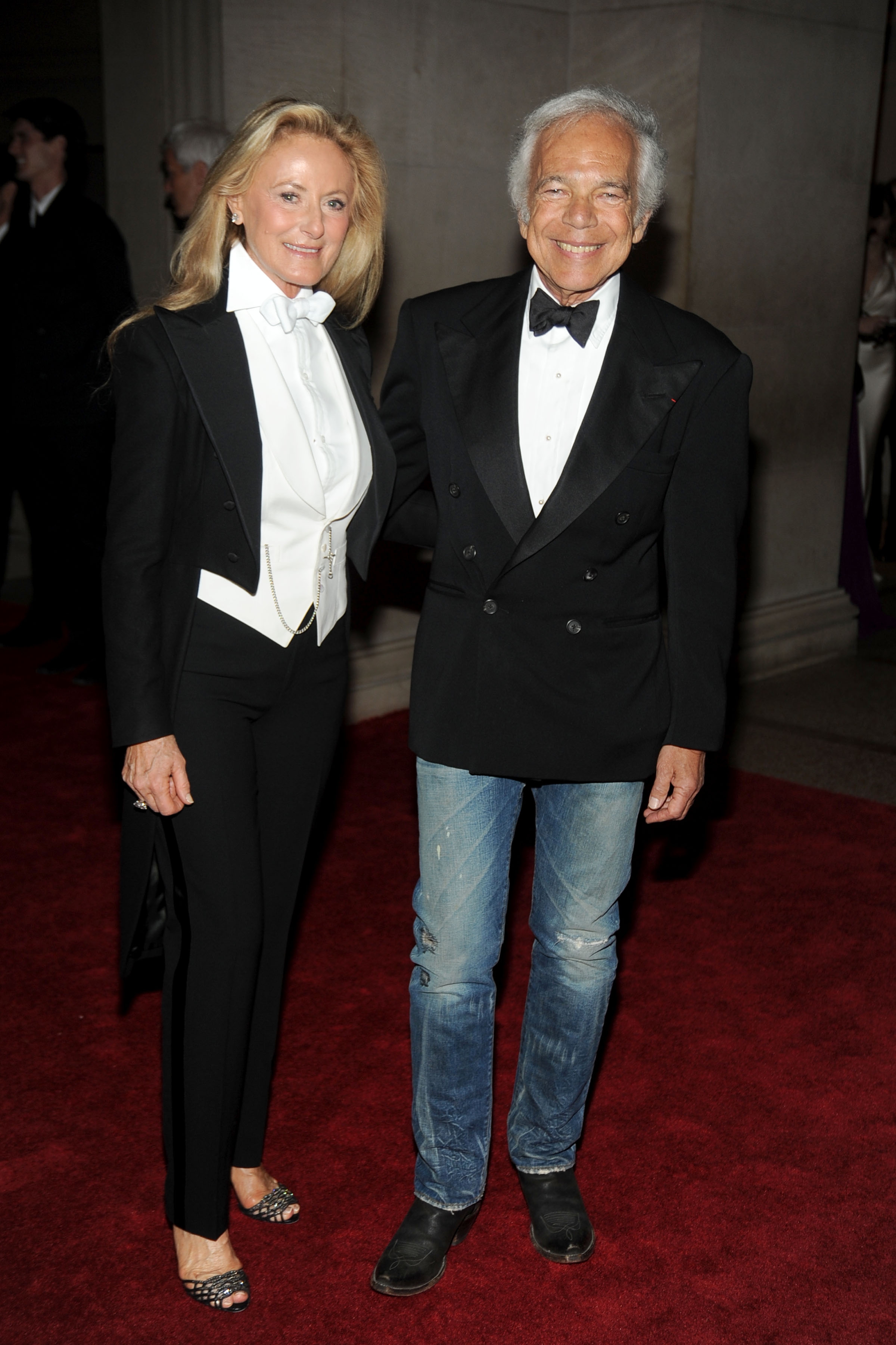 Two people on a red carpet; one wears a tuxedo jacket with a bow tie, and the other wears a double-breasted jacket with jeans and bow tie