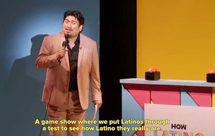 A person on a game show stage in a suit, holding a microphone, standing near a table with buzzers. Subtitles mention a test for how Latino they really are