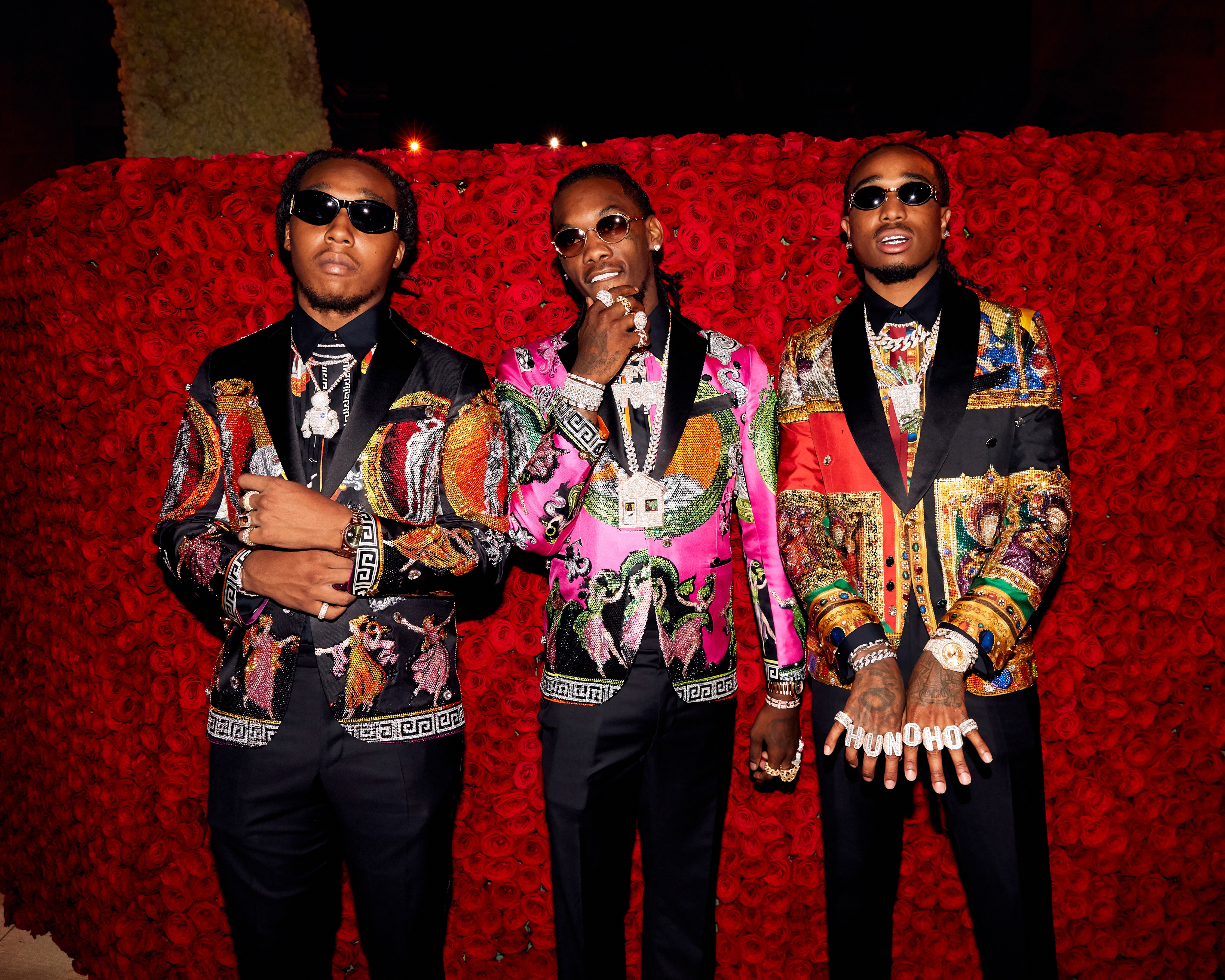 Three individuals in ornate, detailed jackets pose in front of a backdrop of roses. The style is bold and luxurious, showcasing intricate patterns