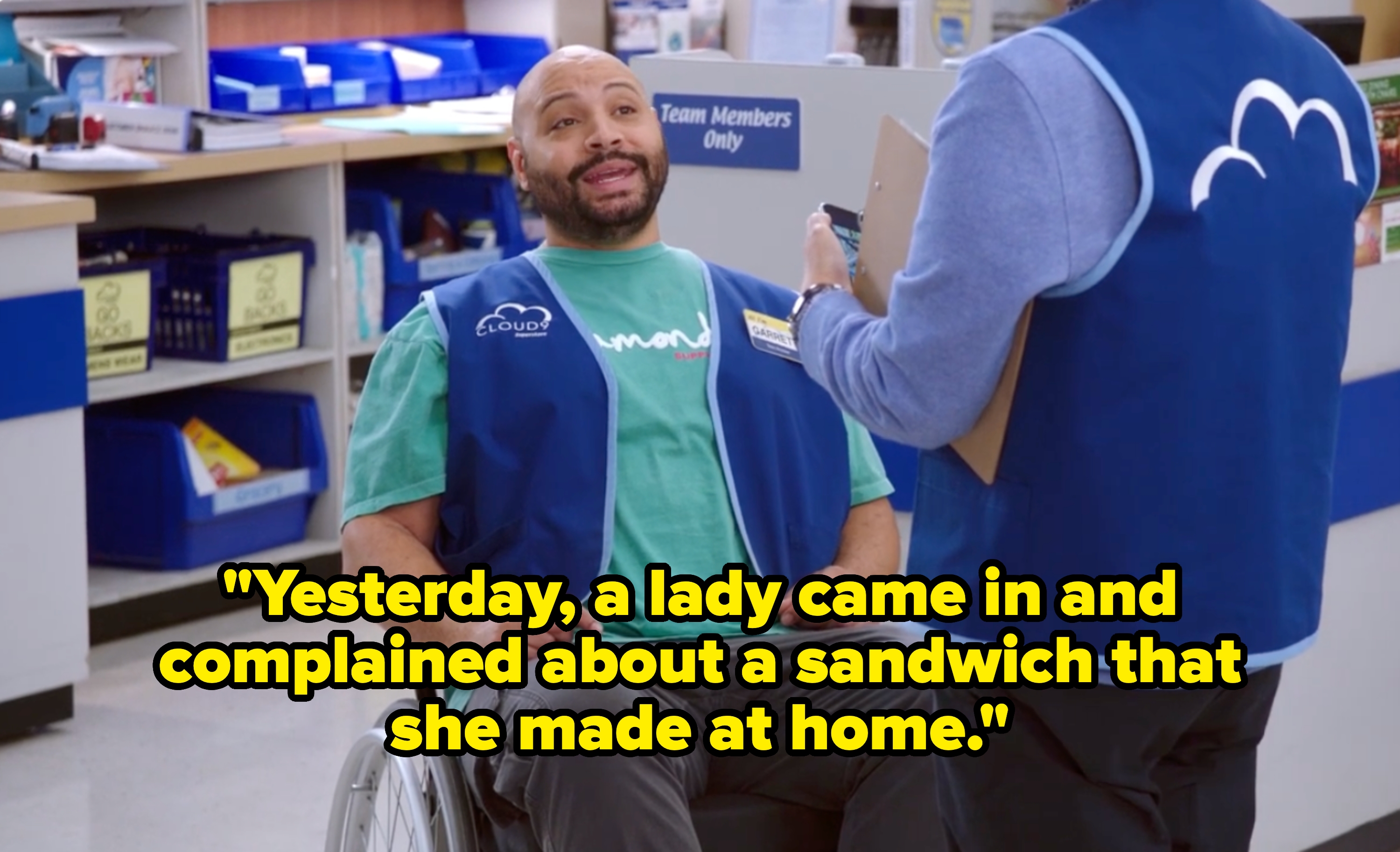 A man in a wheelchair wearing a &quot;Cloud 9&quot; vest is talking to a coworker with a clipboard in a retail store setting