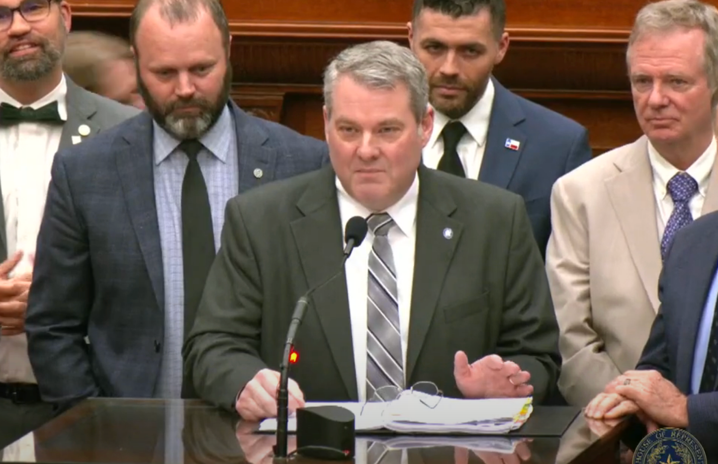 A group of suited men, one speaking at a podium with a microphone, looks serious. They appear to be at an official event or press conference