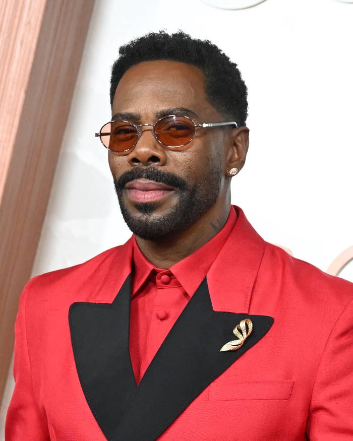 Celebrity in a red carpet setting wears a striking red suit with a black lapel and brooch, complemented by round sunglasses