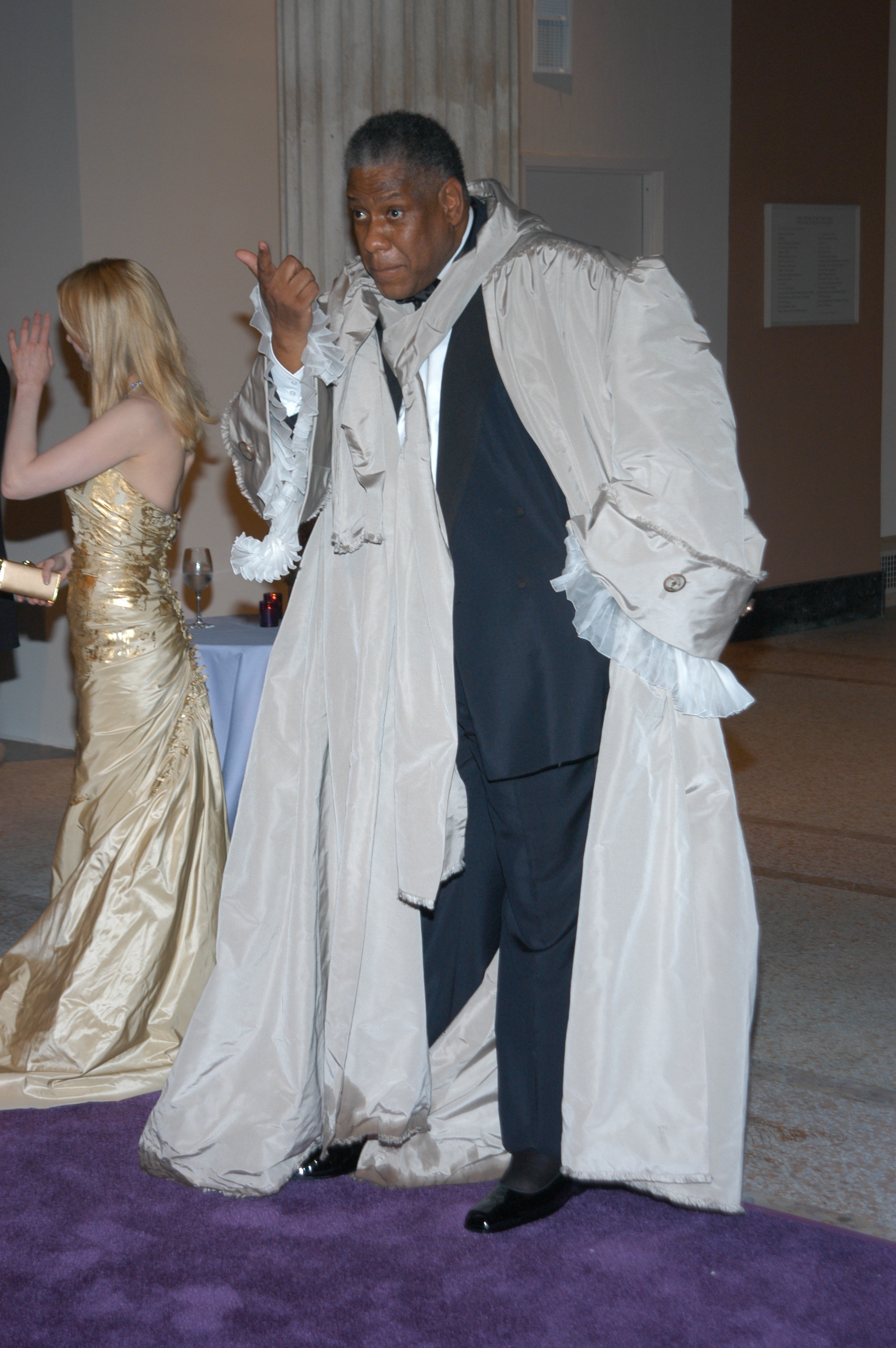 Person in a dramatic, oversized coat and scarf on a red carpet, standing next to someone in an elegant evening gown