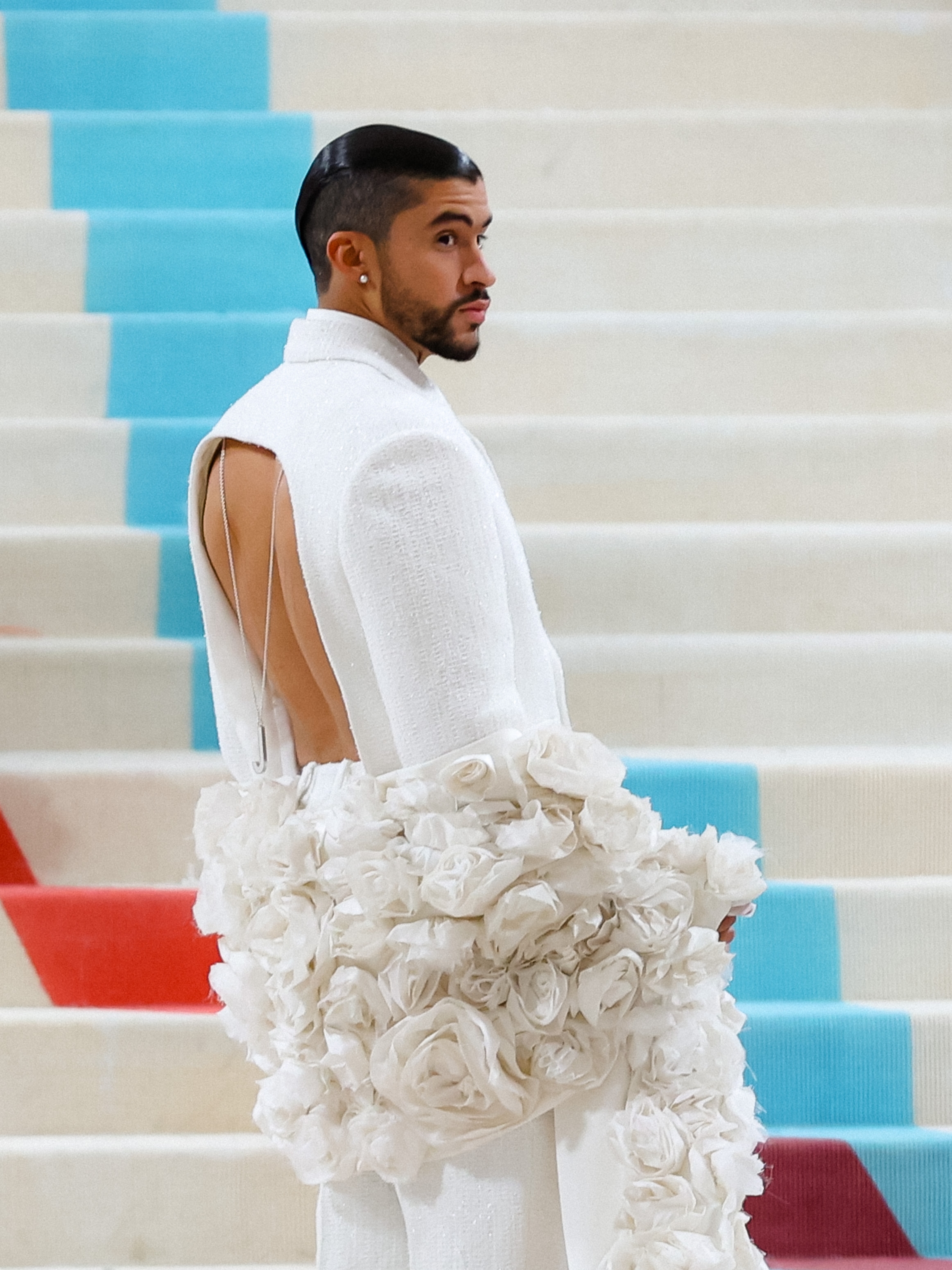 Person on carpeted steps wearing a fashionable open-backed white outfit adorned with large, sculptural rose-like fabric details